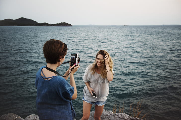 short haired woman in blue top taking photo of another woman in white top standing beside body of water