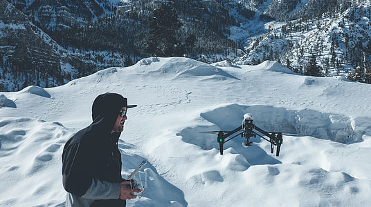 man wearing pull-over jacket infront of black quadcopter on snow field mountain
