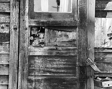 grayscale photo of girl and boy hiding behind wooden door