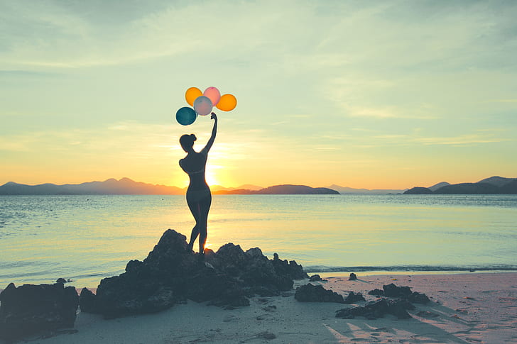 silhouette photo of woman holding assorted-color balloons standing on rock formation during sunset