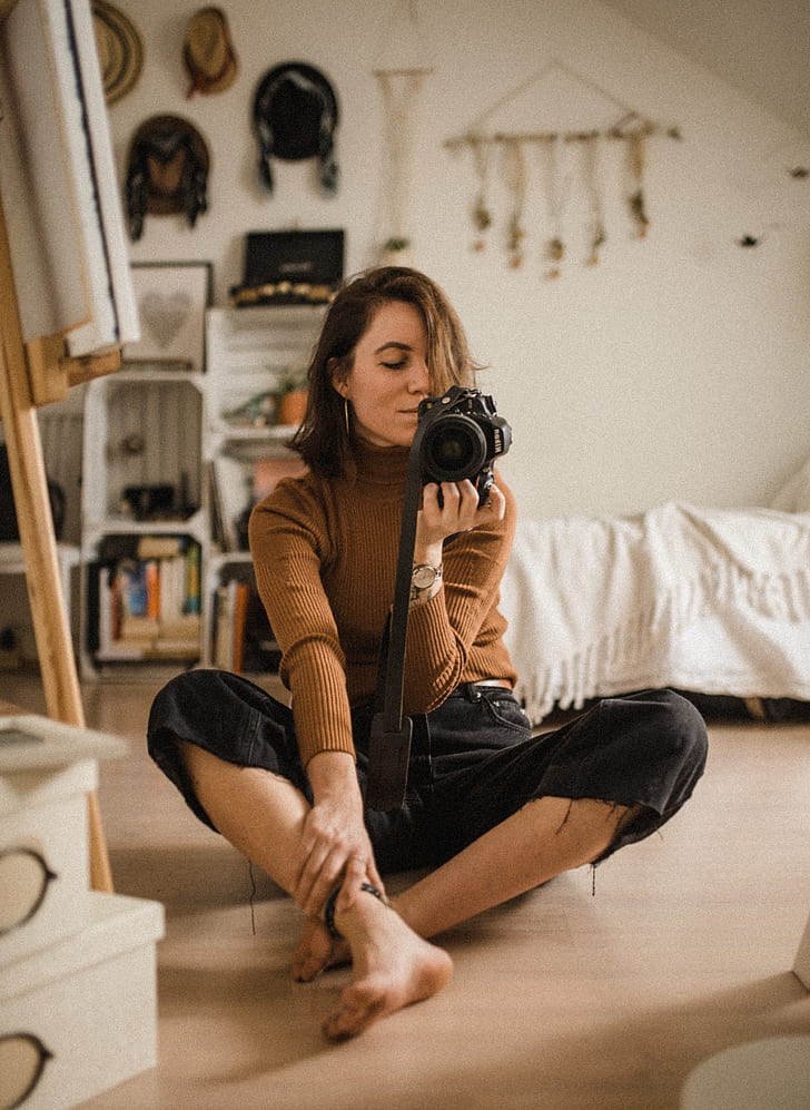 focus photo of woman in brown sweater and black bottoms holding black DSLR camera