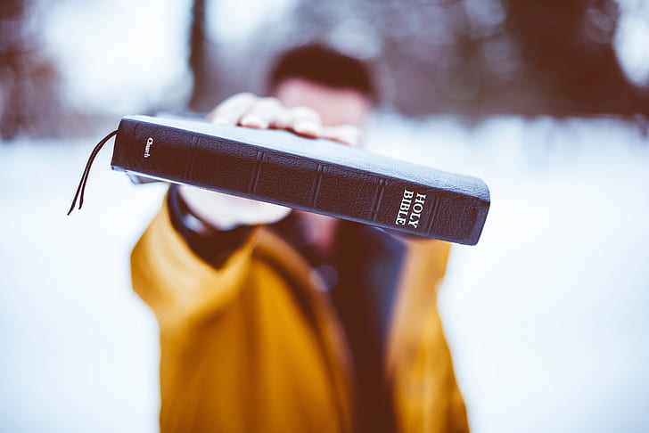 selective focus photography of man wearing brown jacket holding brown Holy Bible