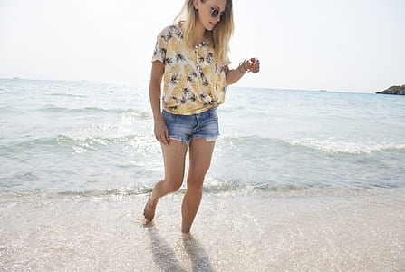 photo of woman wearing brown, gray ,and black floral top and blue short shorts walking in seashore