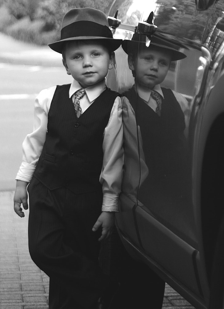 grayscale photography of boy in dress shirt with vest and fedora hat leaning on car