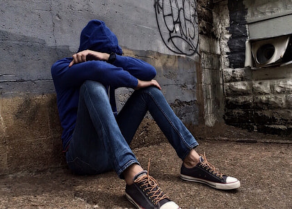 person in blue pullover hoodie and blue jeans sitting while leaning on gray concrete wall during daytime