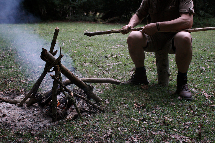 man sitting on log making fire with wood pile during daytime