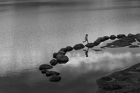 grayscale photography of person hopping on stones in body of water