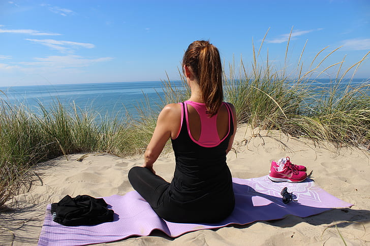 woman in black sleeveless top and black leggings sitting of purple yoga mat on sand near body of water during daytime