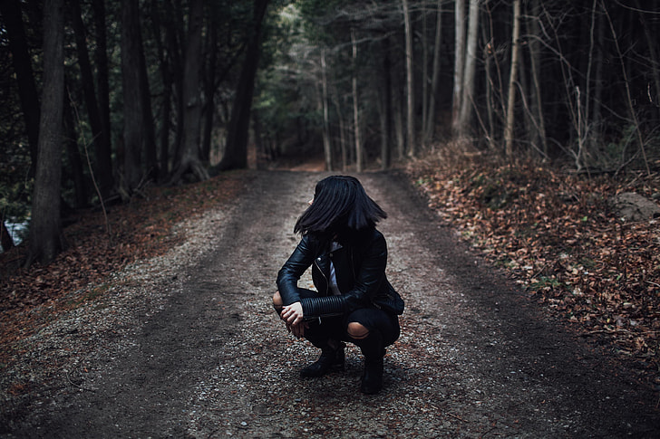 woman in black leather jacket siting on dirt road between trees during daytime