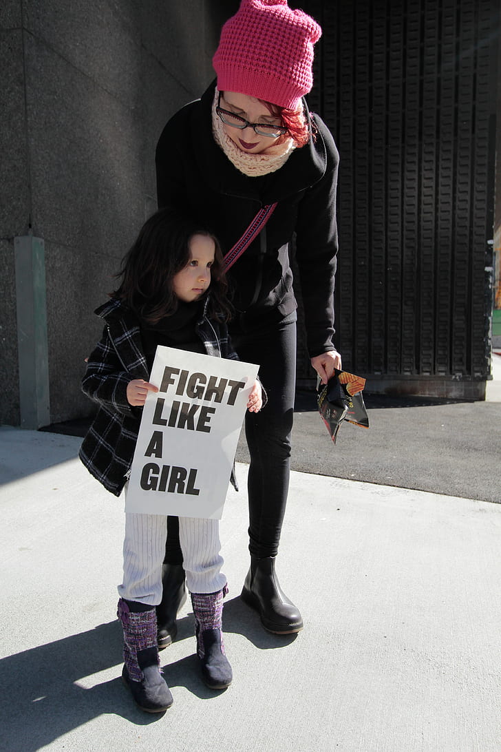 girl in black and white plaid jacket holding white fight like a girl-printed signage during daytime