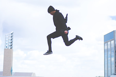 photography of man wearing black beanie, black top, black pants, and black low top sneakers jumping under gray sky
