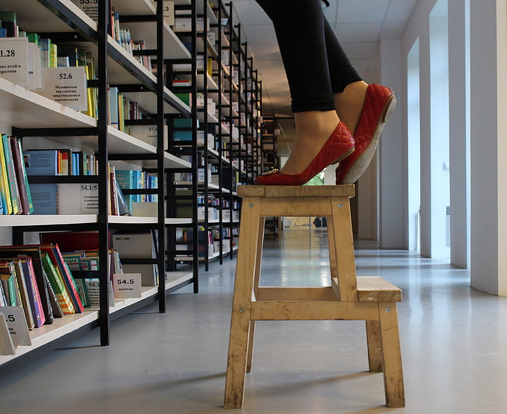 person in red shoes standing on wooden step ladder inside library