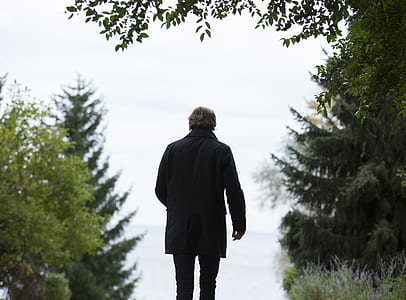 Shallow Focus Photography of Man Wearing Black Coat and Black Pants Standing Beside Green Trees