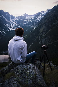 Man in Gray Hoodie Sitting on Gray Stone With Dslr Camera Tripod