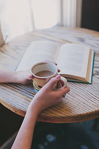 woman holding half-filled coffee cup and book on table