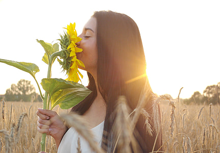 woman wearing white top holding yellow sunflower while standing on brown field during daytime