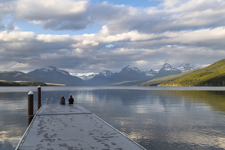 two person sitting on black wooden dock while looking at mountain with body of water scenery during daytime