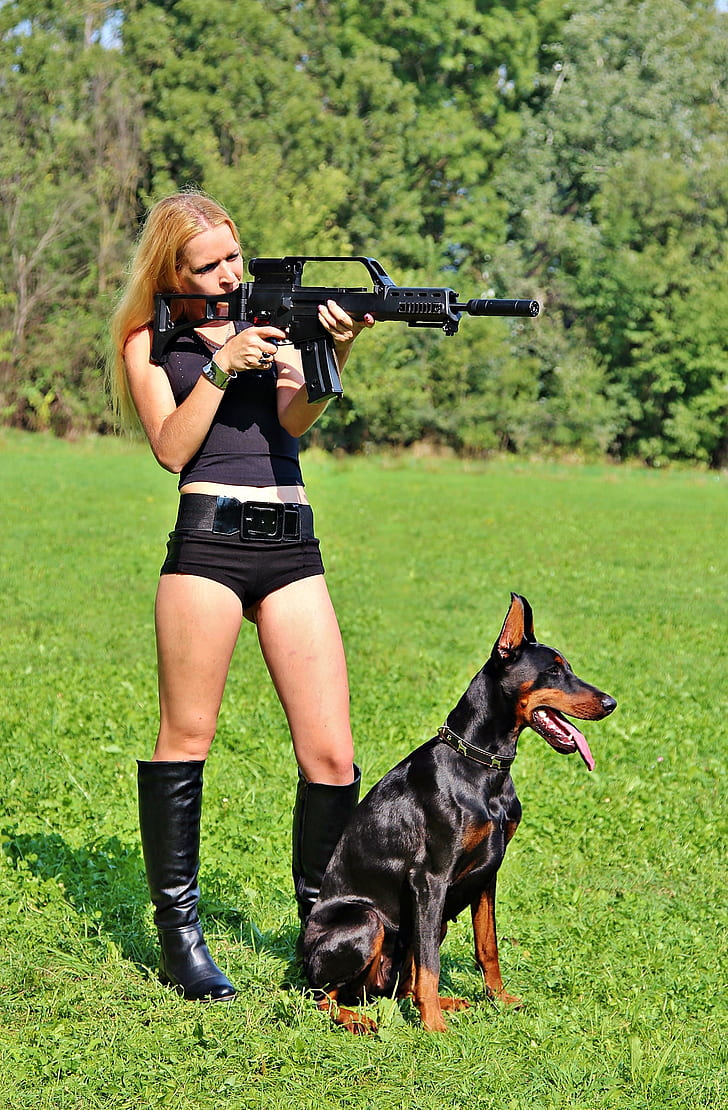woman holding a rifle with black and tan Doberman pinscher during daytime