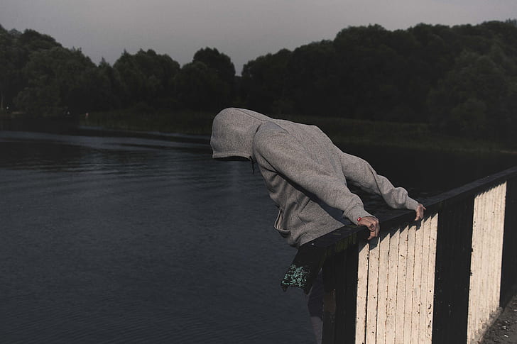 person wearing grey pullover hoodie holding on brown and white metal railings overlooking body of water