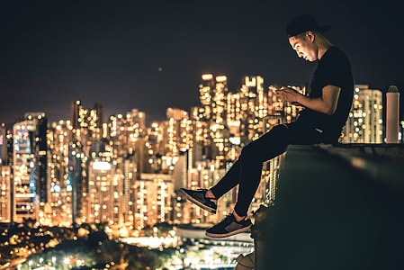 photo of man in black cap, T-shirt, pants, and low top sneakers sitting on edge with high rise buildings with lights during night time