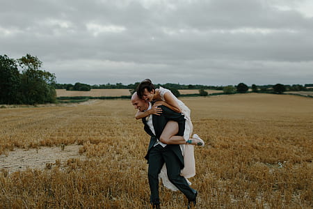 woman piggy back riding man in wearing black suit on brown grass field under cloudy sky