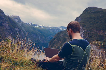 man holding laptop sitting on grass field facing valley with river