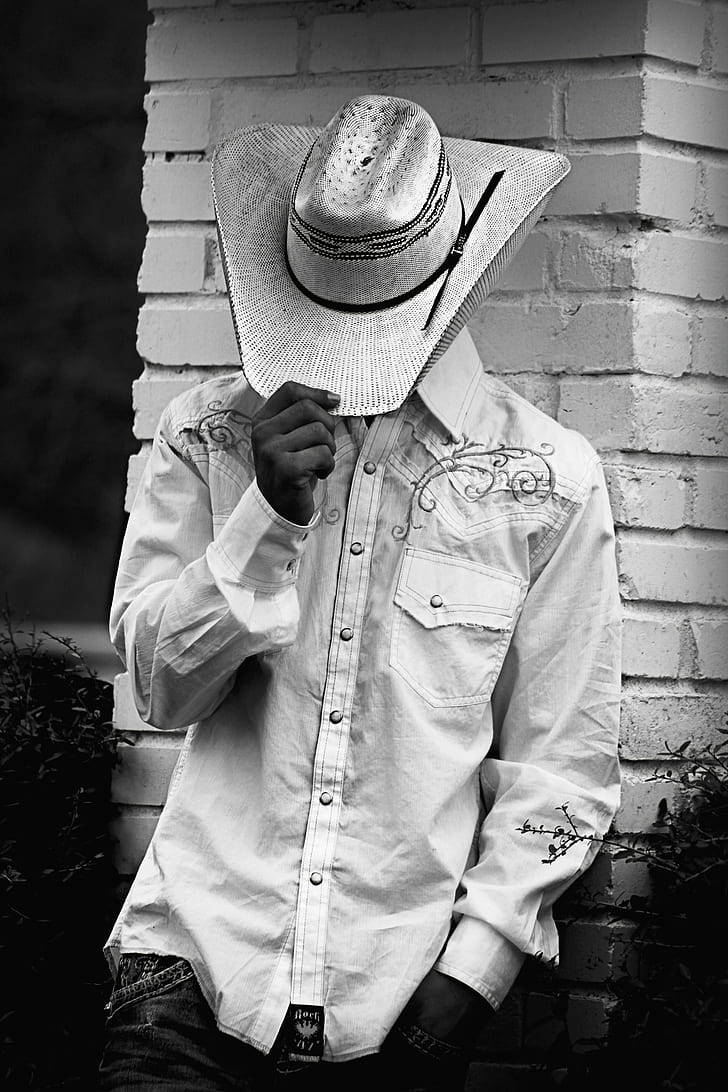 grayscale photography of man wearing floral dress shirt and cowboy hat leaning on bricked wall