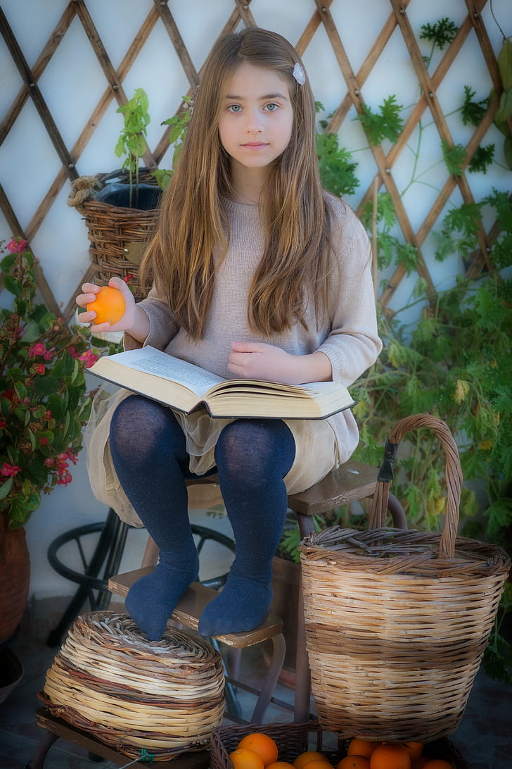 girl in gray knit sweater holding book and orange fruit sitting on chair beside brown wicker basket
