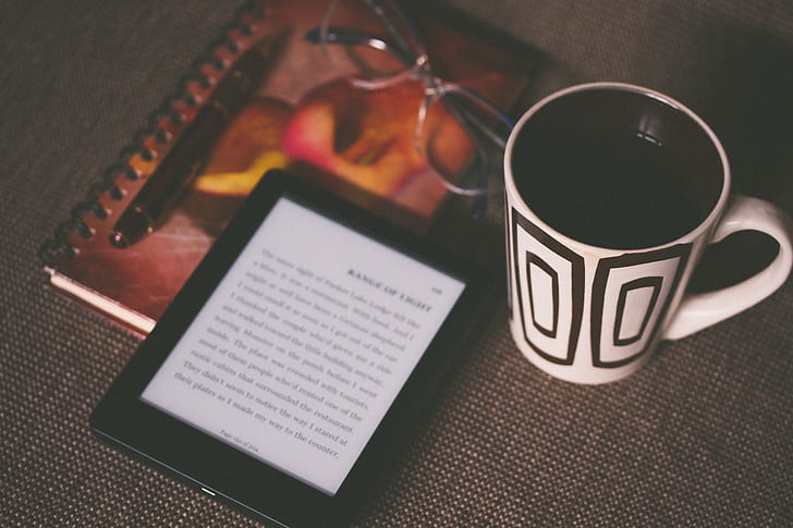 white and black ceramic mug beside black Amazon Kindle ebook reader on gray surface