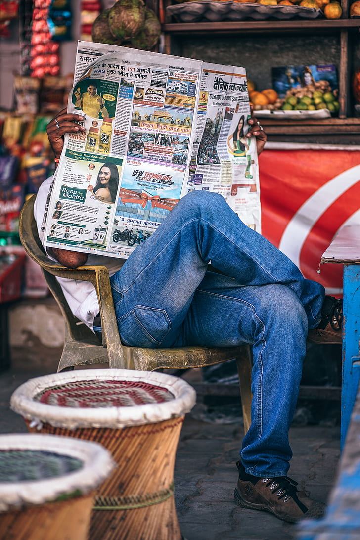 tilt shift lens photography of man wearing blue jeans while holding newspaper