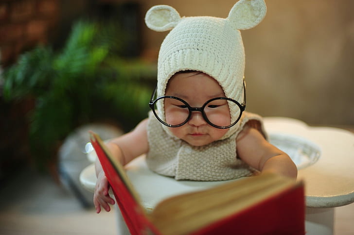 baby wearing gray knitted pram suit and eyeglasses holding book
