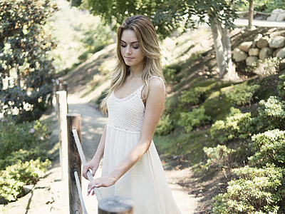 selective focus photography of woman in white dress holding on brown wooden fence while looking down
