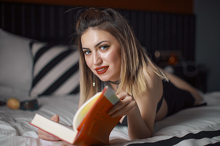 woman lying on bed reading book while smiling