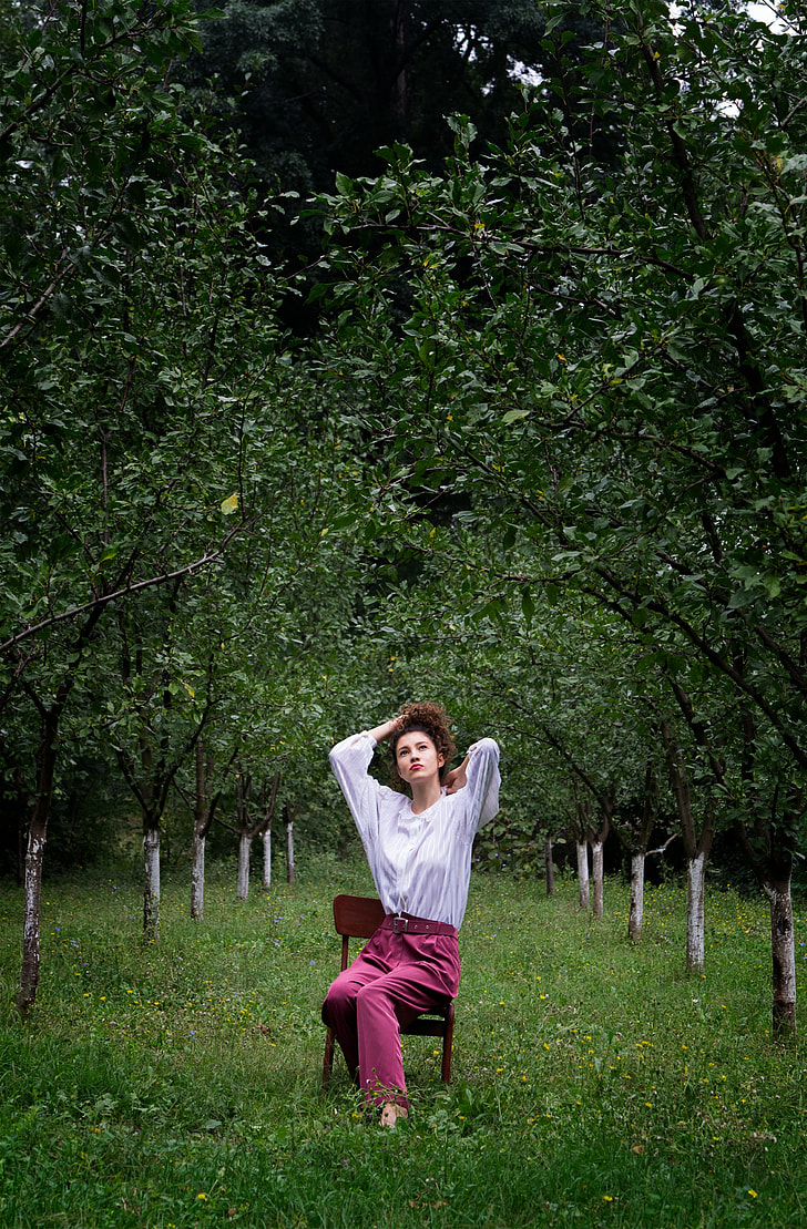 photo of woman in white long-sleeve top with red pants sitting on brown chair surround trees