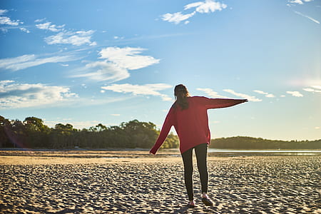 Woman in Red Sweater and Black Pants on White Sand Near Body of Water During Daytime
