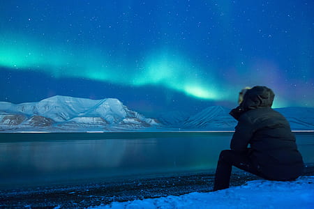 person wearing black ski jacket sitting in front of body of water with distance at snowy hills under dark sky with Aurora borealis