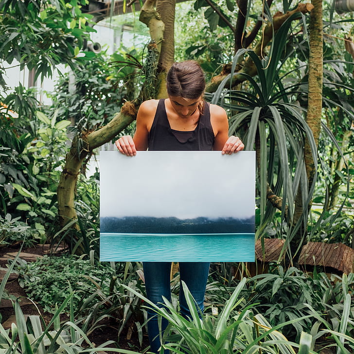 person wearing black tank top holding poster with body of water near tree print