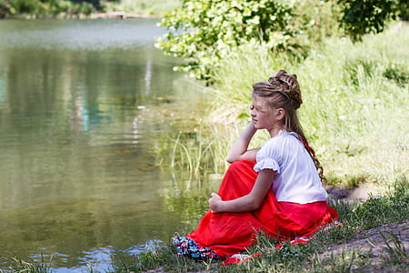 woman wearing white blouse and red maxi skirt sitting on green grass beside body of water