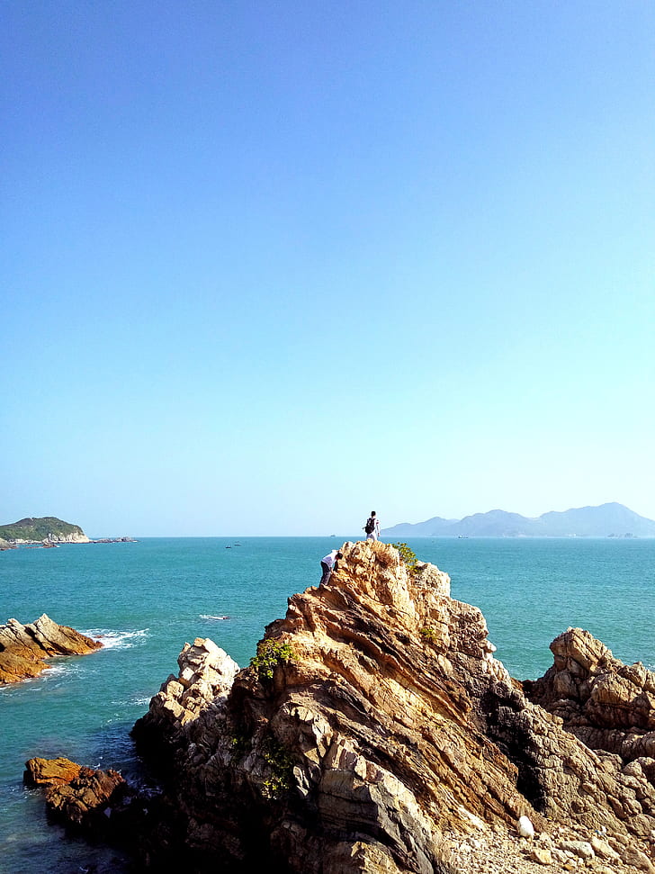 person on top of brown and yellow stone formation overlooking the sea under blue sky during day time