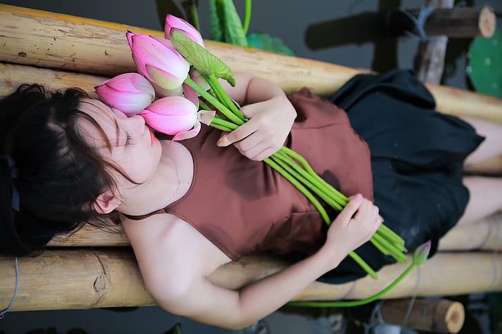 woman holding pink petaled flowers while lying on bamboo