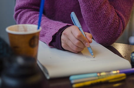 selective focus photography of person holding pen writing on paper on table near coffee cup