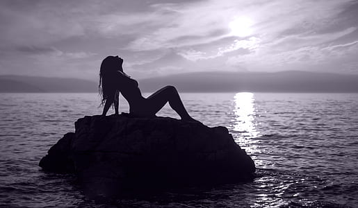 silhouette photography of woman sitting on rock formation near body of water