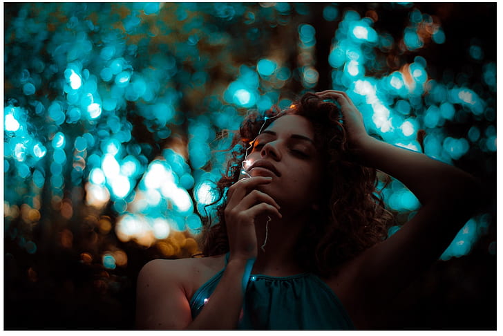 selective focus photography of woman in teal sleeveless top holding white string lights with bokeh brackground