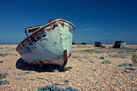 An old abandoned boat lies on a shingle beach on a sunny day. Image captured in Dungeness, Kent, England