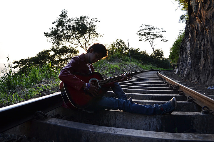 man playing a guitar sitting on train rail at daytime