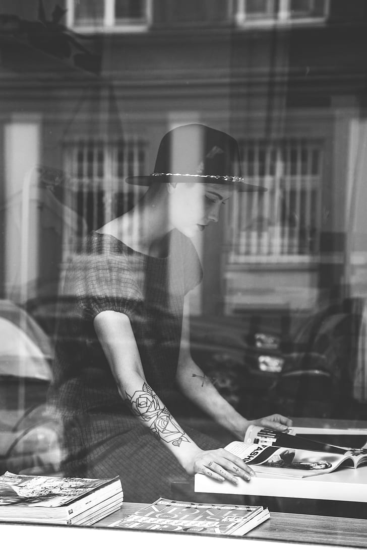 grayscale photo of woman wearing black short-sleeved dress and black hat reading magazine