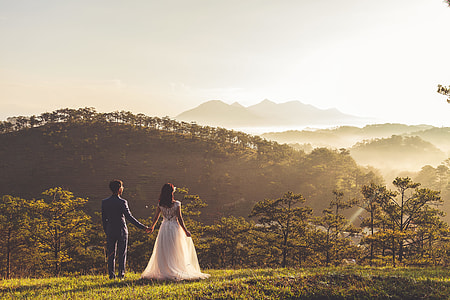 silhouette photo of groom and bride on green field near mountain under cloudy skies during daytime