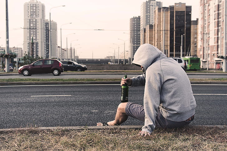 man wearing hoodie sitting beside the road holding bottle near cars during day