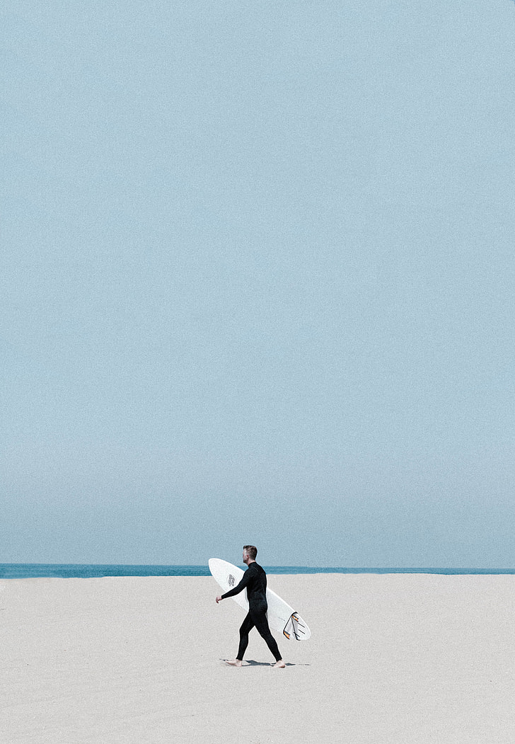 man wearing black wetsuit holding white surfboard walking on shoreline