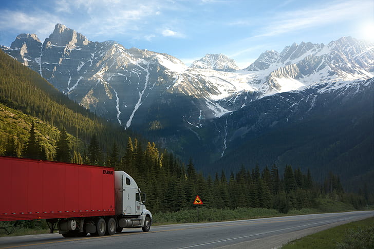 white and red trailer truck running on open road with snow-capped mountains background during daytime